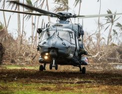 A Royal New Zealand Air Force NH90 helicopter lands on the grass in Fiji, palm trees blowing the background as well as mess from Cyclone Winston