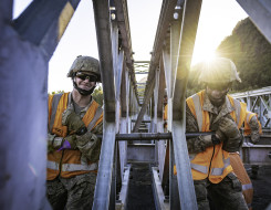Two New Zealand Army soldiers lift part of a Bailey Bridge, one on each side of it. The image is backlit with sun and has been taken in the centre of the bridge.