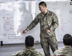 A New Zealand Army soldier points a whiteboard with content on it. In the foreground, blurred are other uniformed people watching on. 