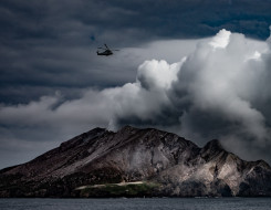 A SH-2G(I) Seasprite Helicopter flying above a thick plume of white smoke coming from Whaakari/White Island following the eruption.