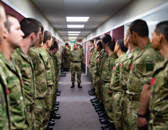 New Zealand Army soldiers stand in a corridor a row of people on each side. One solider stands in the middle of the corridor. 