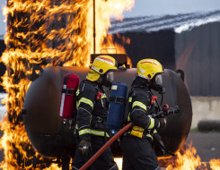 Two NZDF firefighters during a training activity. Both are in full breathing apparatus and one is carrying a hose. There are flames around them.