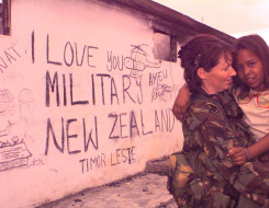 A New Zealand Army soldiers holds a young child in the right of the image. On the left is the wall of a building that reads "I love you military New Zealand Timor Leste - Ameu' with military drawings around it. The photo has a pink colouring to it. 