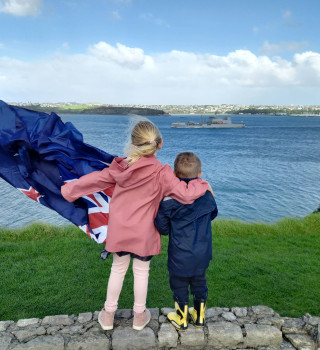 Family members wave farewell to Royal New Zealand Navy's HMNZS Aotearoa in Auckland. The two children are holding a flag and one of them has their arm around the other. 