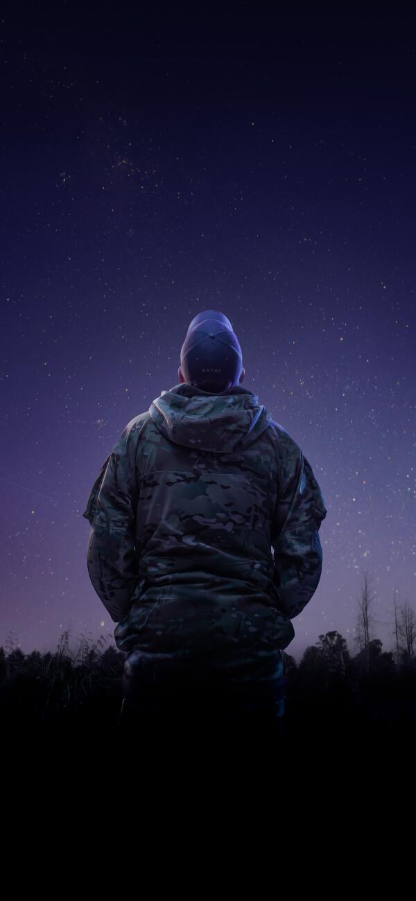 An aviator from the Royal New Zealand Air Force looks to the stars while wearing a camouflage jacket and a cap that has the letters 'RNZAF' on the back. 