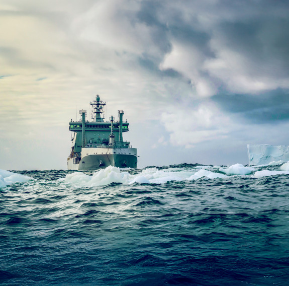 HMNZS Aotearoa seen bow on in grey green Antarctic seas which meet grey and cloudy skies about halfway. Light sea ice floats ahead of the ship and a distant flat topped, square angled ice shelf projects into the scene from the right.