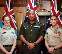Image of three uniformed personnel standing in a line, in front of flags. All are looking at the camera smiling slightly with their hand together in front of them. 