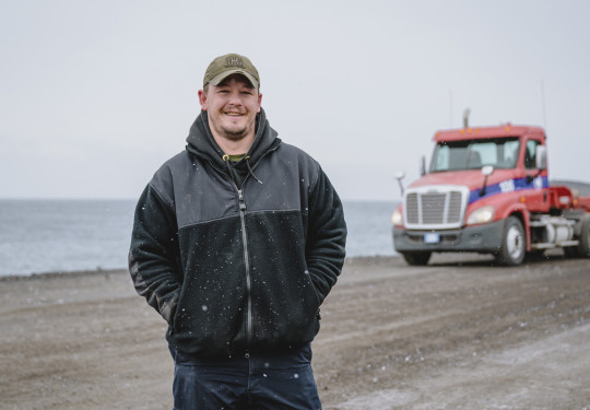 A person stands in the snow, looking and smiling at the camera. Hands in pockets and wearing a peaked cap. There is a truck in the background, along with some water.