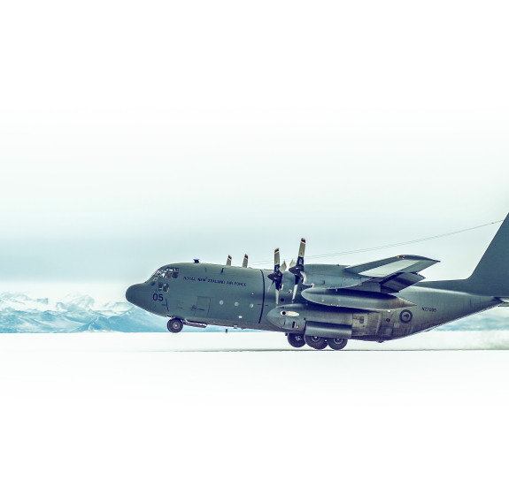 An RNZAF C-130H touches down with its rear under carriage on an ice runway. Under a white sky, a range of snow covered mountains stretch the width of the scene on the horizon.