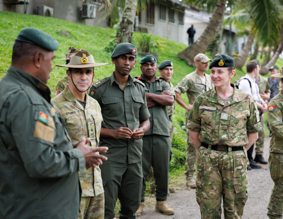 Uniformed military personnel stand outside - all looking at one person to the left of the photo who is speaking. 