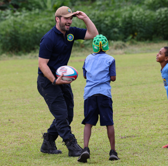 LT Cunningham smiles while holding a rugby ball with local children out on a field.