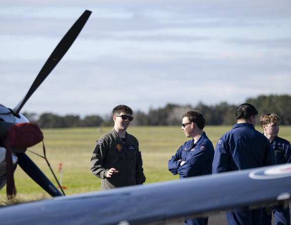 Standing behind an aircraft wing and propellor, an aviator talks with a cadet with an airfield and forest on the horizon.