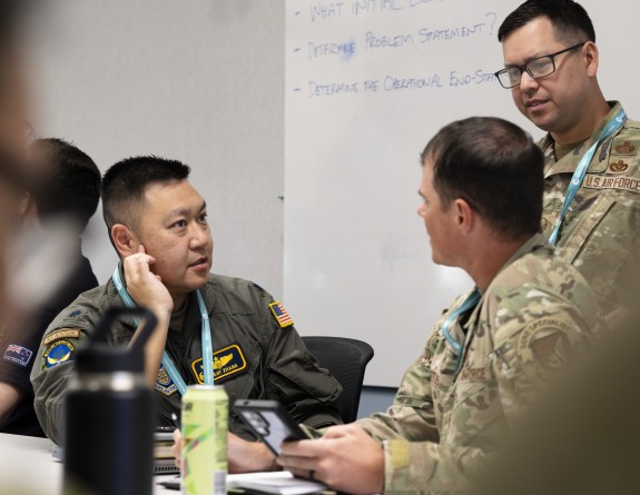 Three military personnel have a discussion in front of a whiteboard, one man is standing while the other two are sitting.