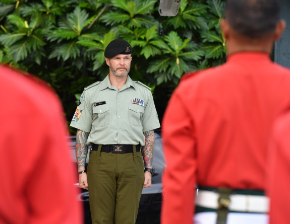 A soldier stands in uniform in between other uniformed soldiers in red. The camera is looking towards the NZ Army soldier but the other soldiers in red have their backs to the camera. 