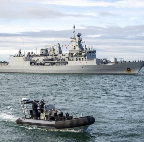 HMNZS Te Kaha sails in with a smaller boat sailing alongside in the foreground. 
